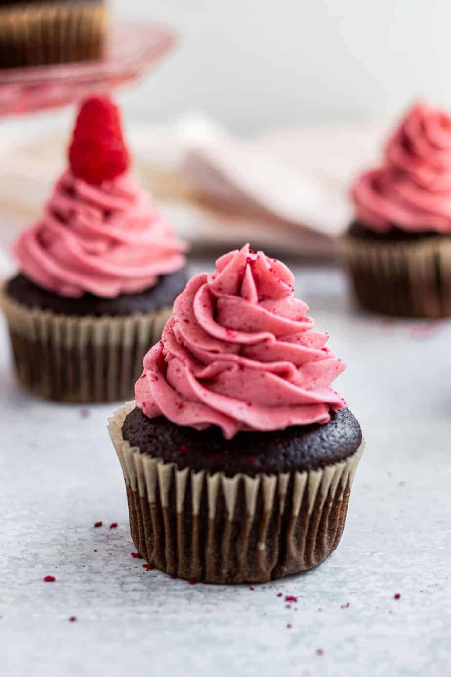 chocolate cupcakes with raspberry buttercream and a pink glass cake stand in the back