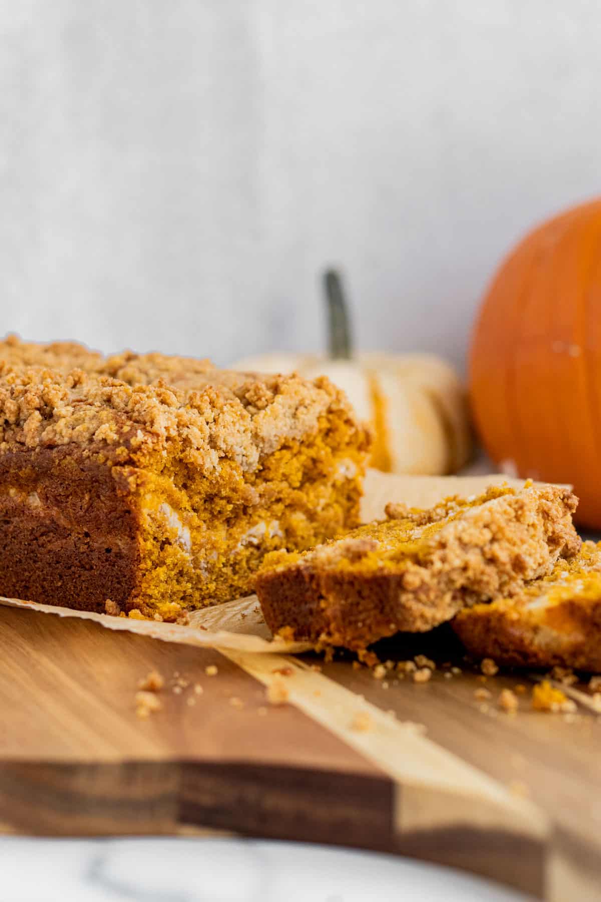 loaf of pumpkin cream cheese bread sitting on a wood cutting board with 2 slices cut. 2 pumpkins in the background.