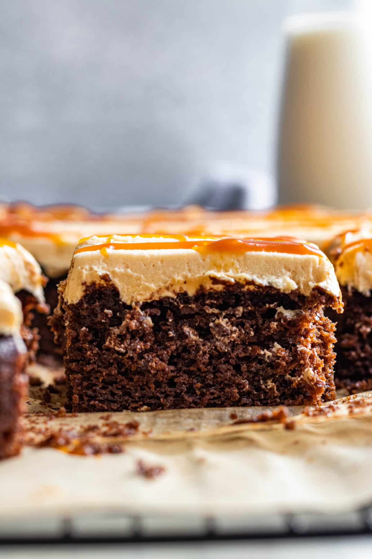 slice of chocolate cake with salted caramel frosting and caramel drizzle on top. Glass of milk in background.