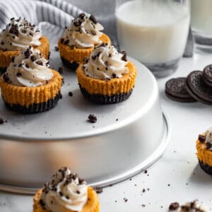 mini pumpkin cheesecakes with whipped cream sitting on silver tin with cookies and milk in the background