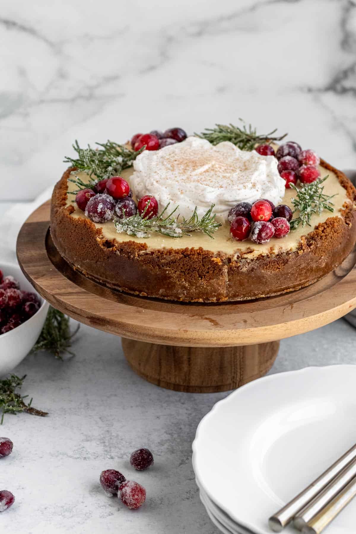 eggnog cheesecake with whipped cream, sugared cranberries and rosemary on a wood cake pedestal with a stack of plates