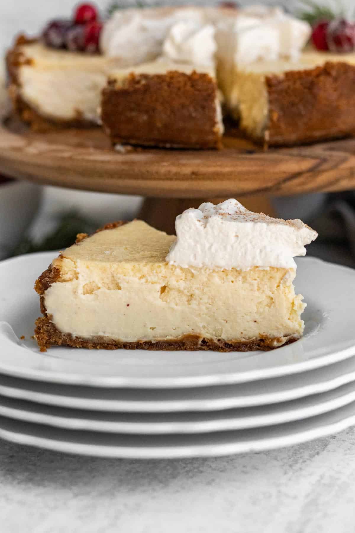 slice of eggnog cheesecake with whipped cream on a stack of white plates with the full cheesecake in the background