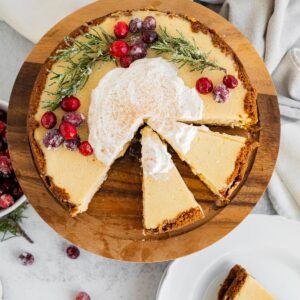 overhead view of an eggnog cheesecake decorated with cranberries and rosemary and 2 slices sliced on a wood cake stand