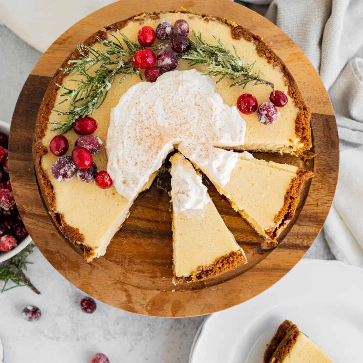 overhead view of an eggnog cheesecake decorated with cranberries and rosemary and 2 slices sliced on a wood cake stand