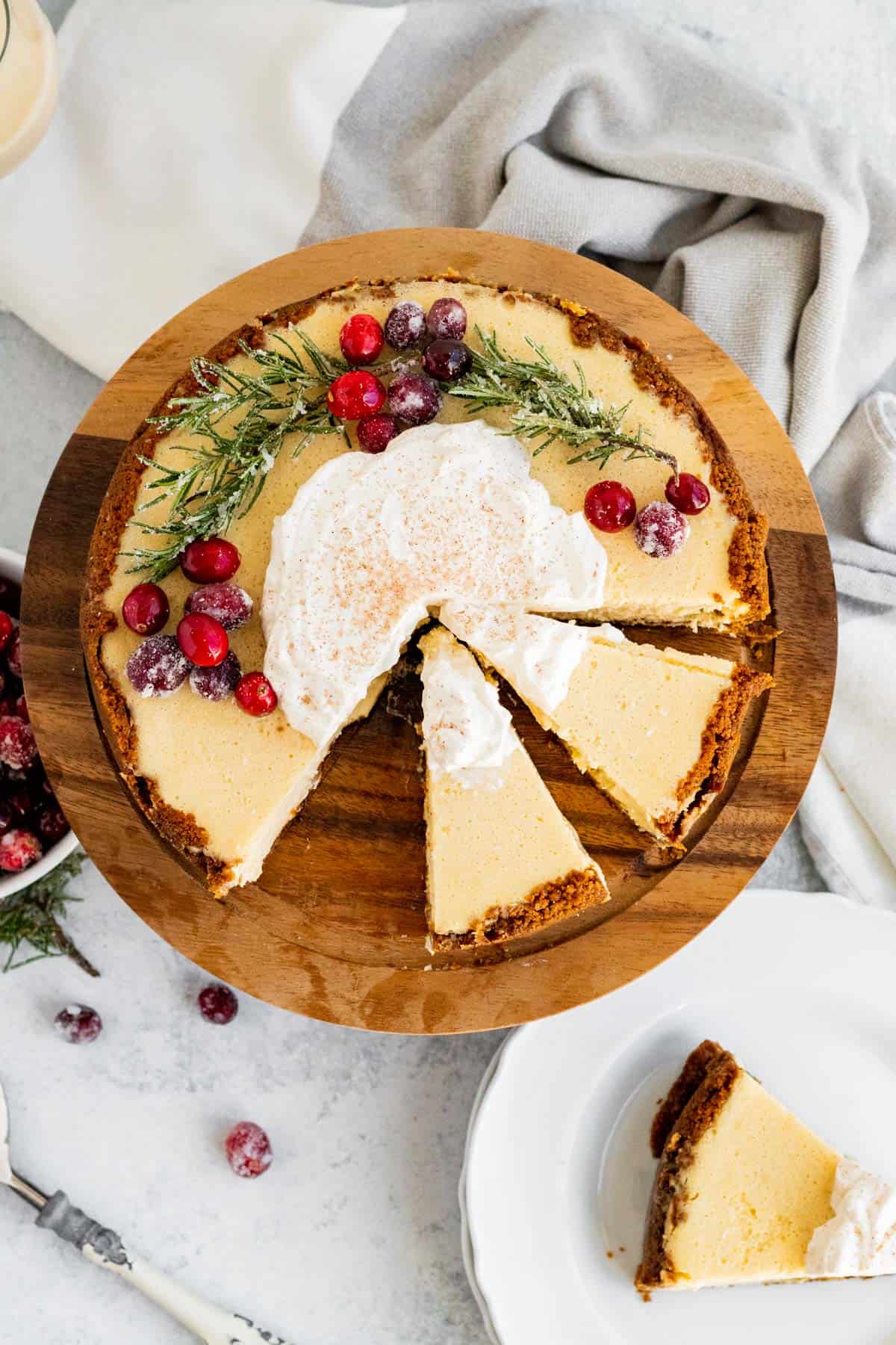 overhead view of an eggnog cheesecake decorated with cranberries and rosemary and 2 slices sliced on a wood cake stand