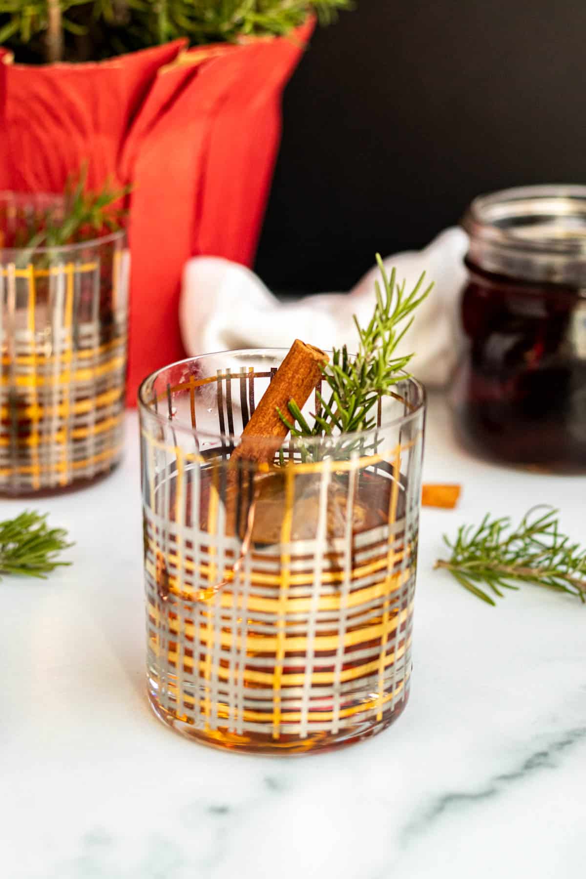 winter old fashioned cocktail in a gold and silver striped rocks glass with rosemary sprigs scattered around 