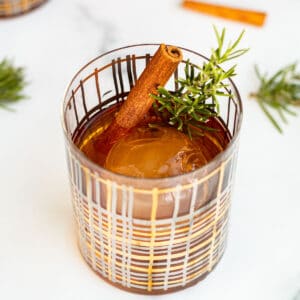 cocktail glass with a clear ice ball and winter old fashioned garnished with rosemary and cinnamon stick sitting on marble counter