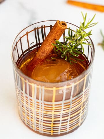 cocktail glass with a clear ice ball and winter old fashioned garnished with rosemary and cinnamon stick sitting on marble counter