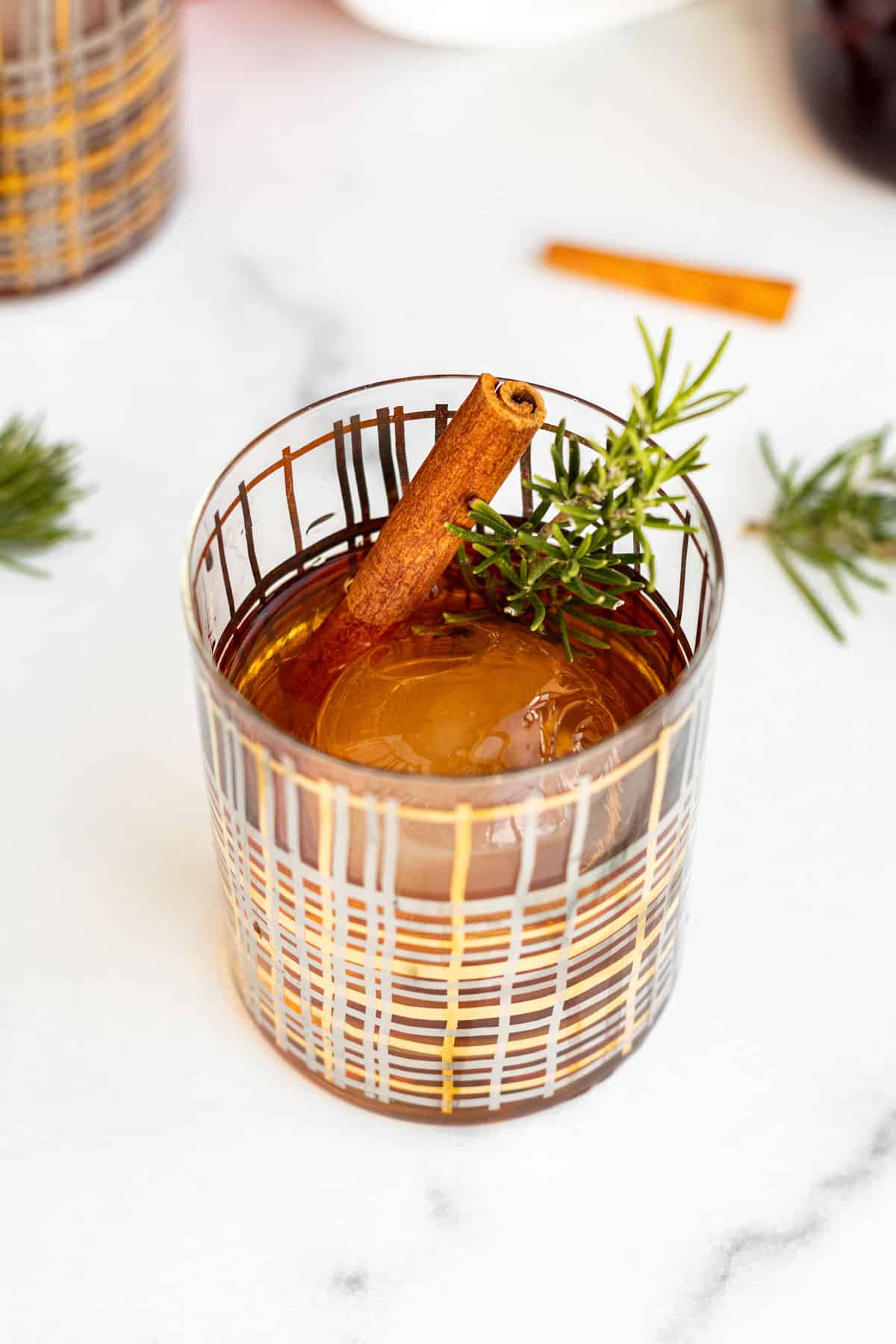 cocktail glass with a clear ice ball and winter old fashioned garnished with rosemary and cinnamon stick sitting on marble counter