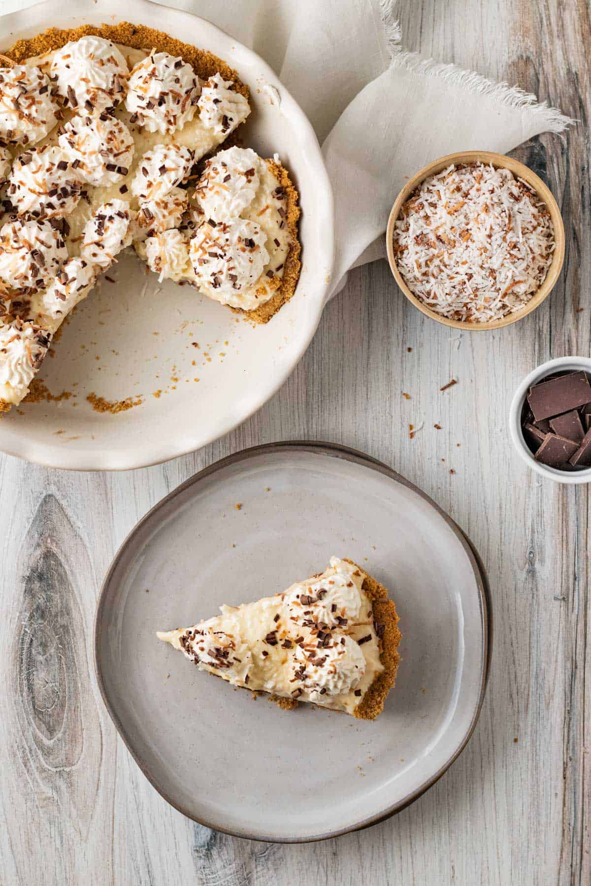 overhead view of chocolate coconut cream pie in white pie dish with slice of pie on a gray plate and small bowls of coconut and chocolate pieces