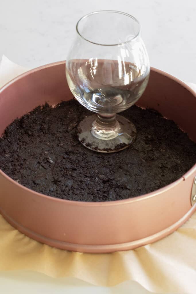 oreo crust being packed down into a pink springform pan with a glass