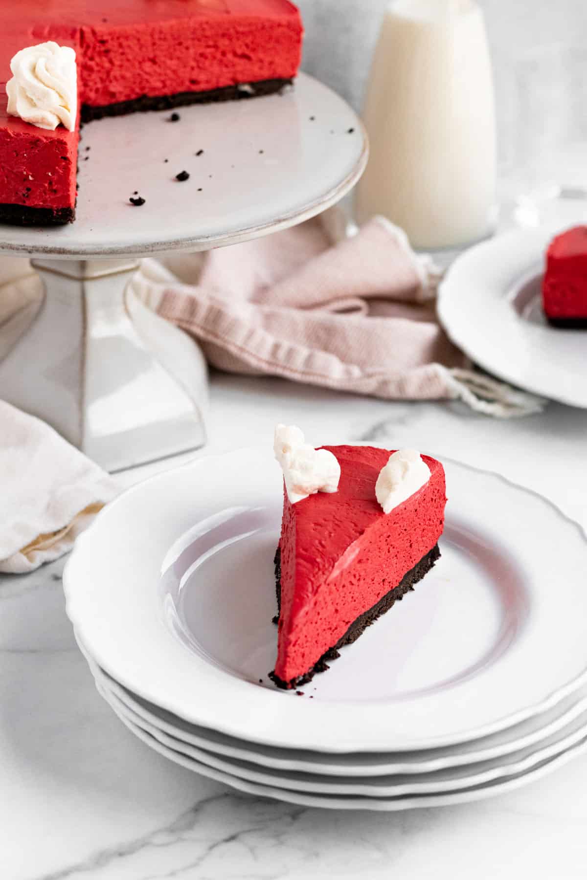 white plate with red velvet no-bake cheescake slice with a pink kitchen towel and glass of milk in the background