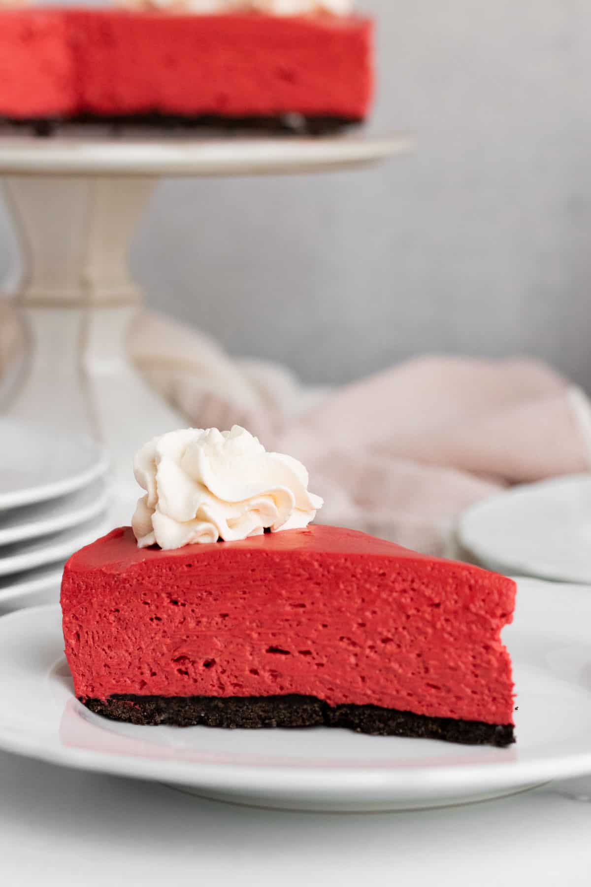 white plate with red velvet no-bake cheescake slice with a pink kitchen towel, stack of white plates and cake stand with the full cheesecake in the background