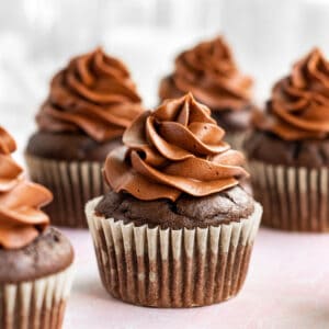 multiple chocolate cupcakes with chocolate buttercream sitting on a pink surface