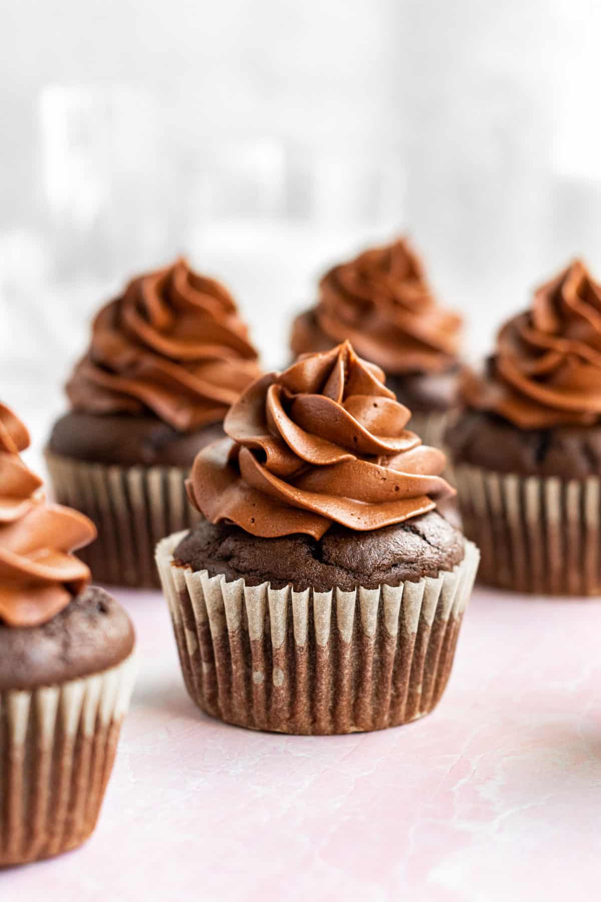 multiple chocolate cupcakes with chocolate buttercream sitting on a pink surface