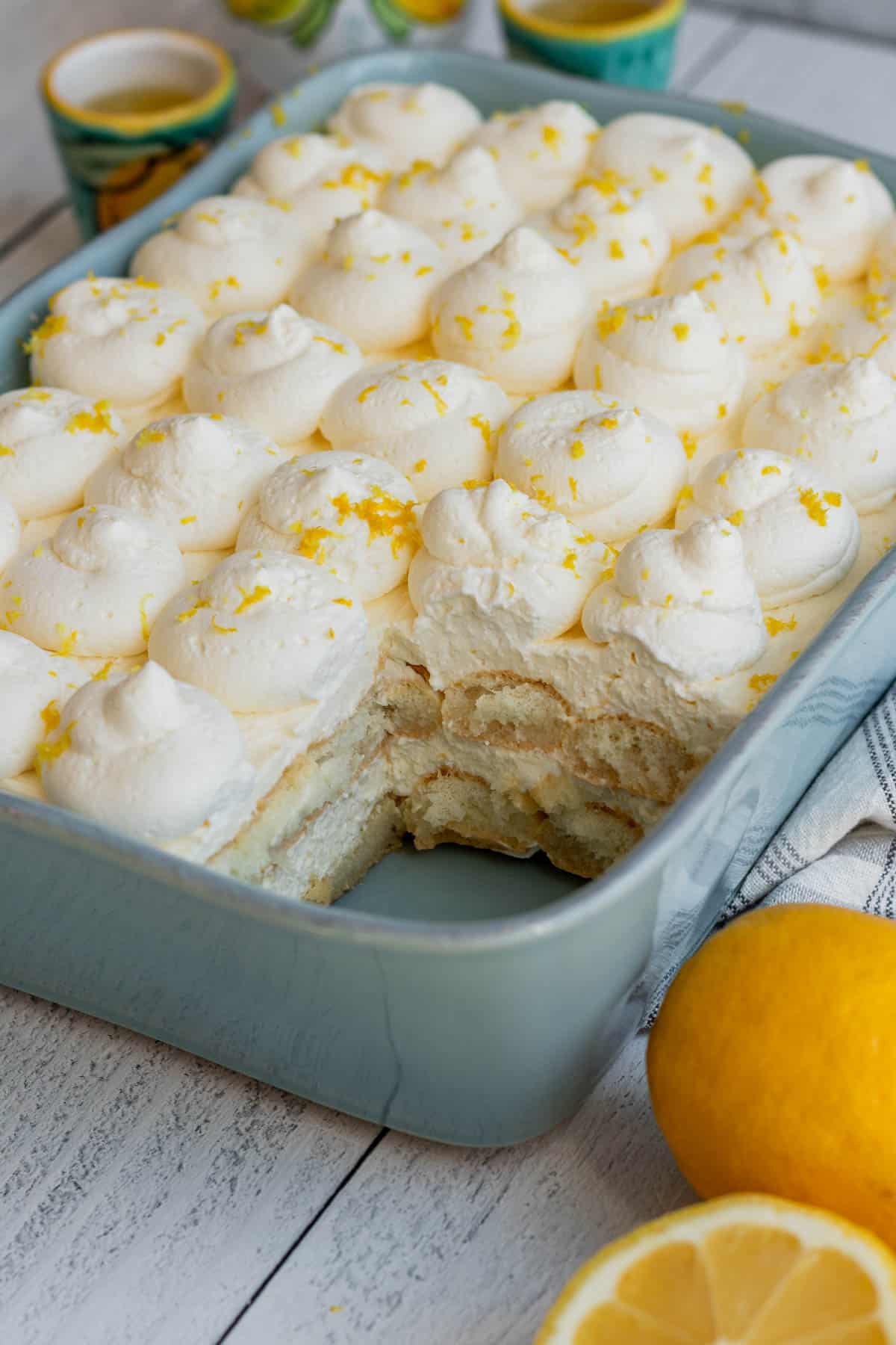 blue baking dish with layers of ladyfingers and lemon mascarpone with lemons to side of baking dish