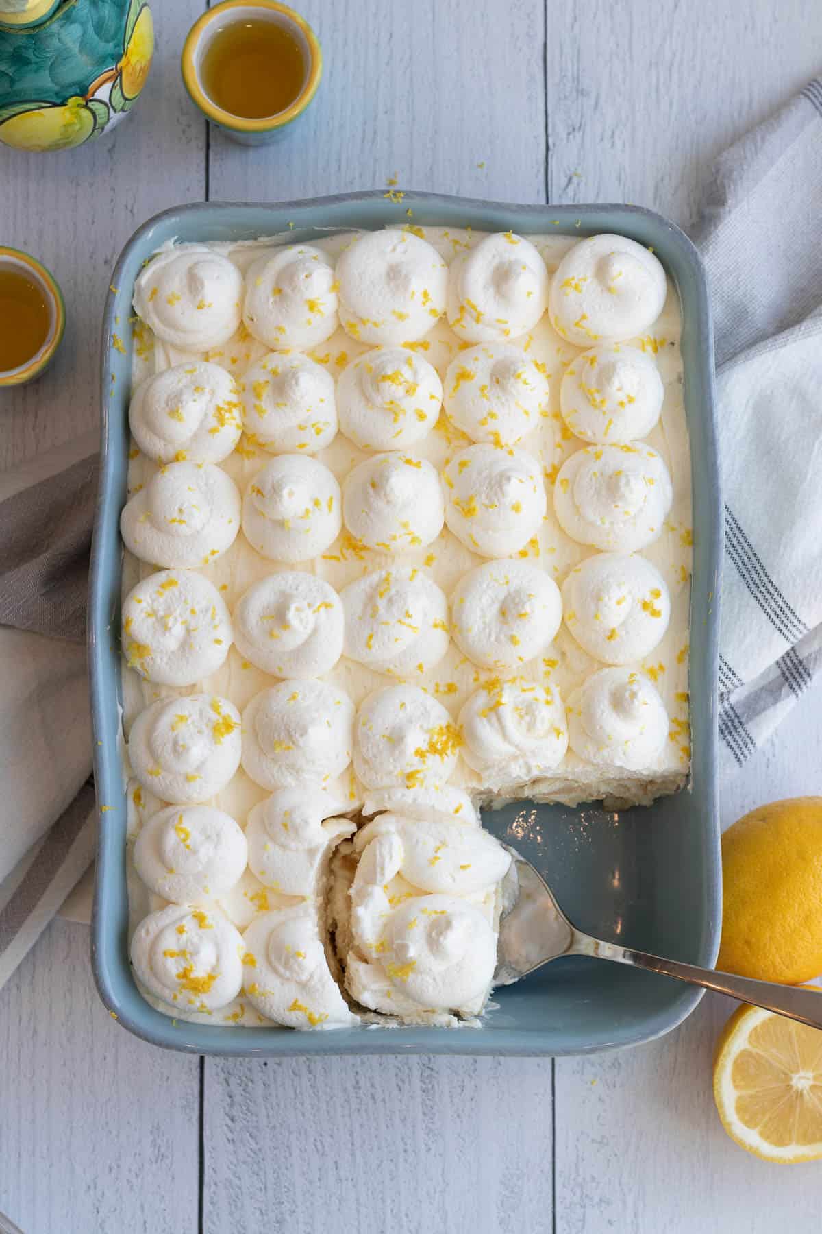 blue baking dish of limoncello tiramisu with a spoon scooping out a serving and lemons and limoncello in the foreground and background