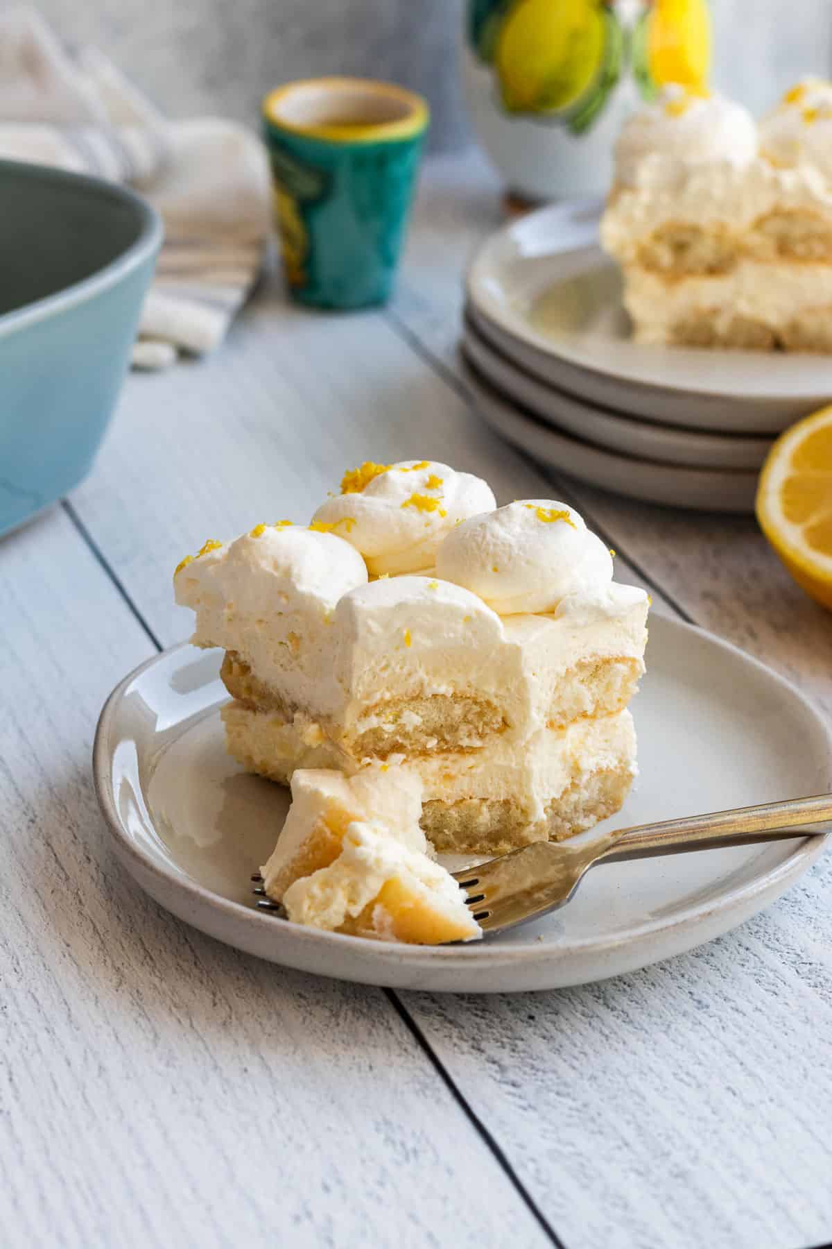 plate of limoncello tiramisu with a gold fork of tiramisu and a stack of plates and another slice of tiramisu in the background