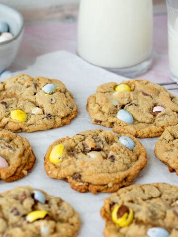 mini egg cookies on parchment paper with glasses of milk in background with bowl of mini egg candies