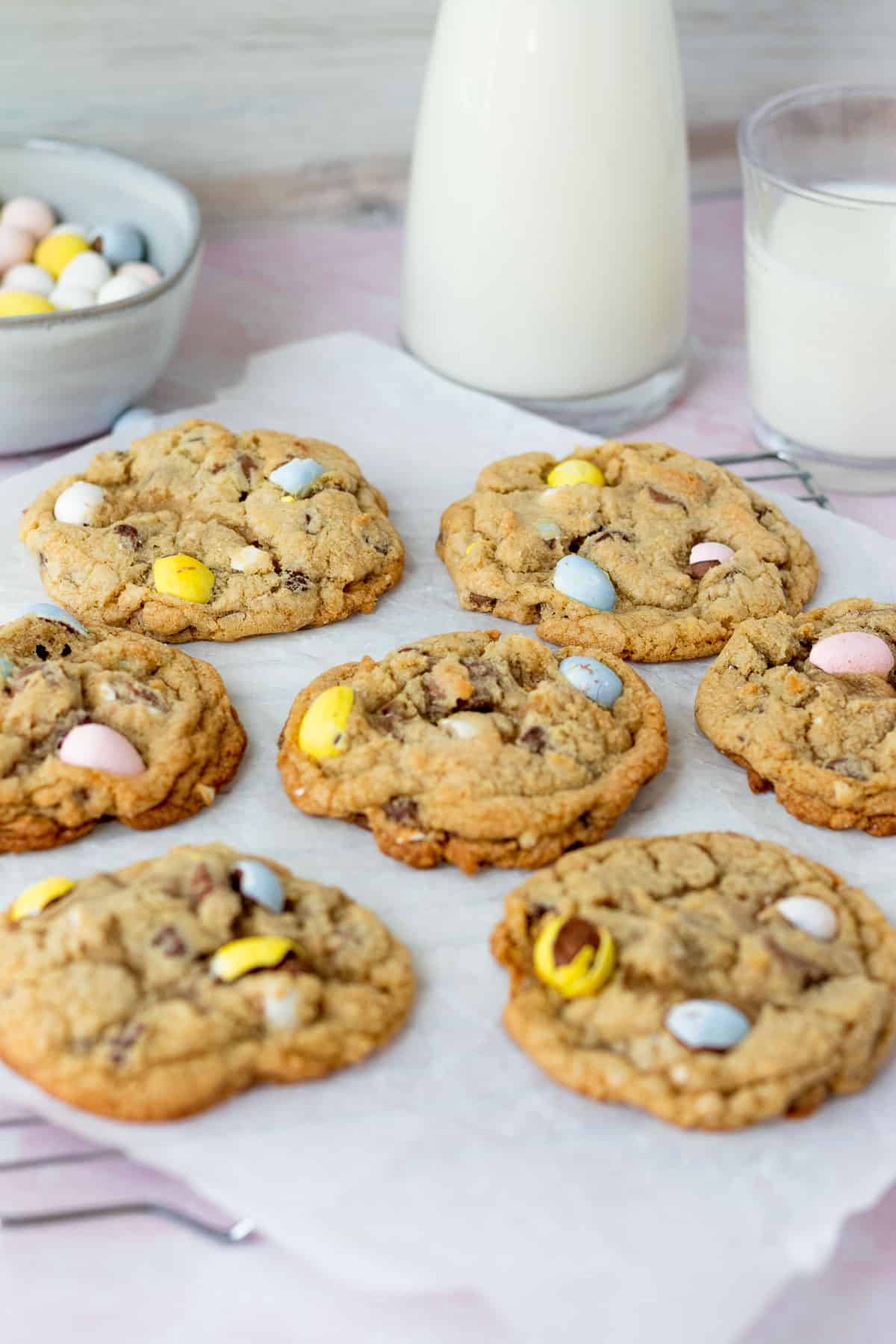 mini egg cookies on parchment paper with glasses of milk in background with bowl of mini egg candies