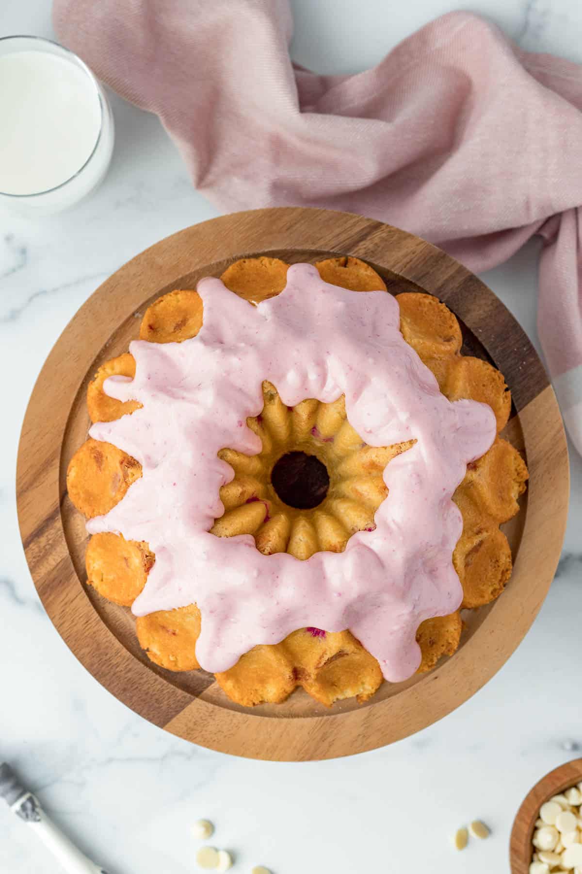 white chocolate raspberry bundt cake on a wood cake stand with raspberry cream cheese glaze and a pink linen