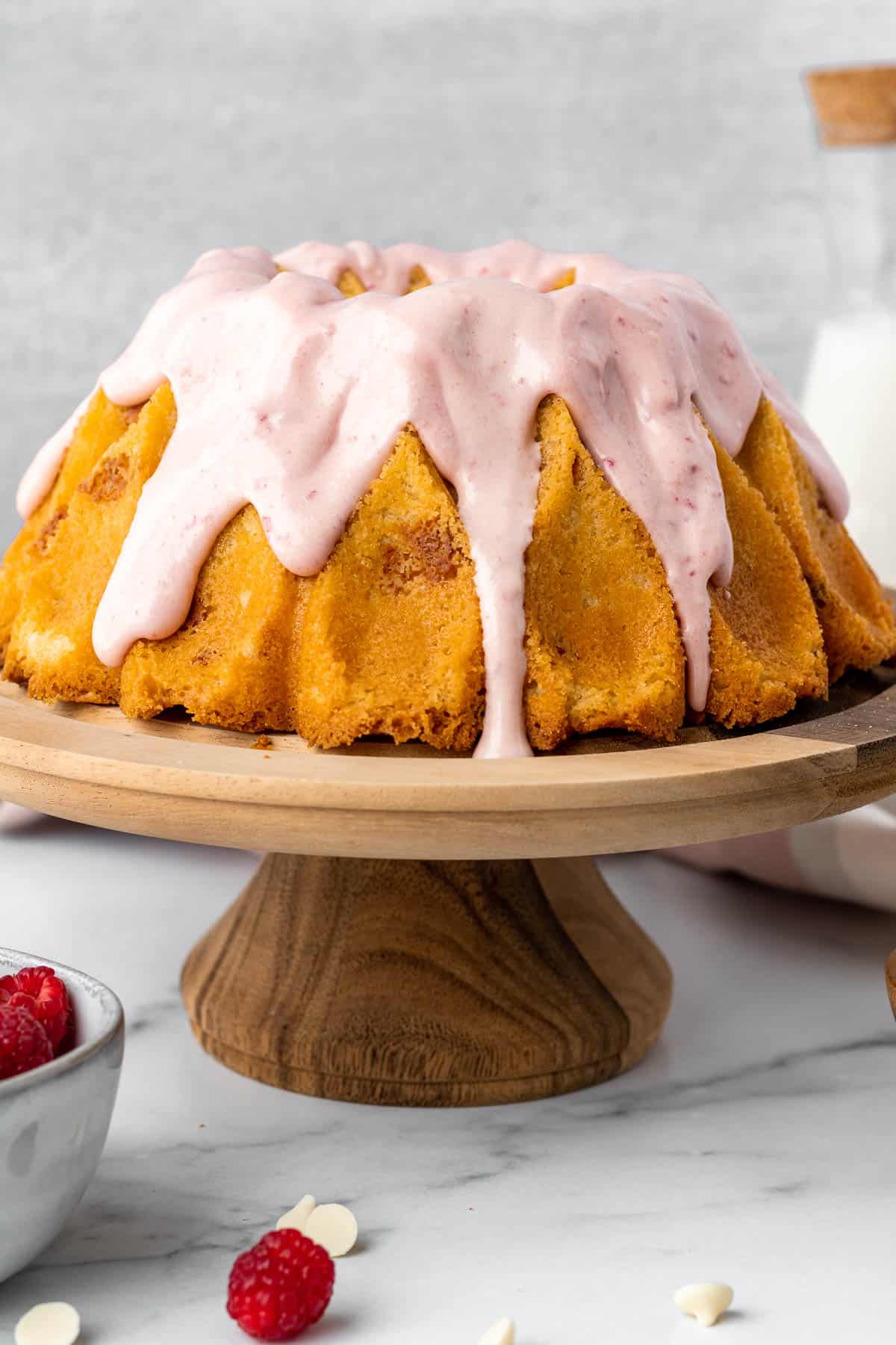 white chocolate raspberry bundt cake on a wood pedestal with bowl of raspberries in foreground and glass of milk in background