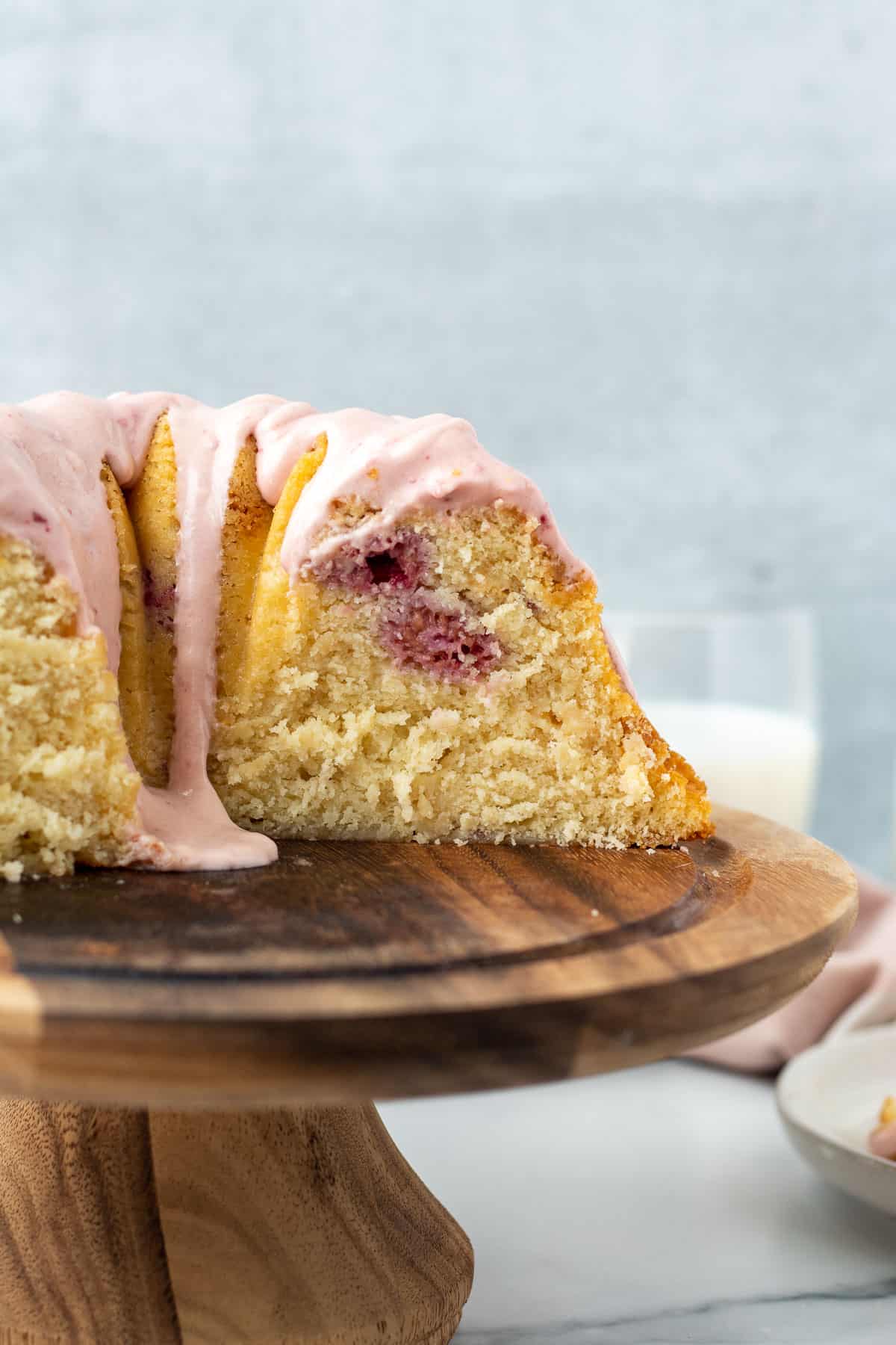 bundt cake with pink glaze on wooden pedestal with slice exposed and glass of milk in background