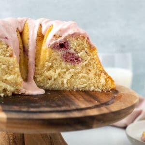 bundt cake with pink glaze on wooden pedestal with slice exposed and glass of milk in background