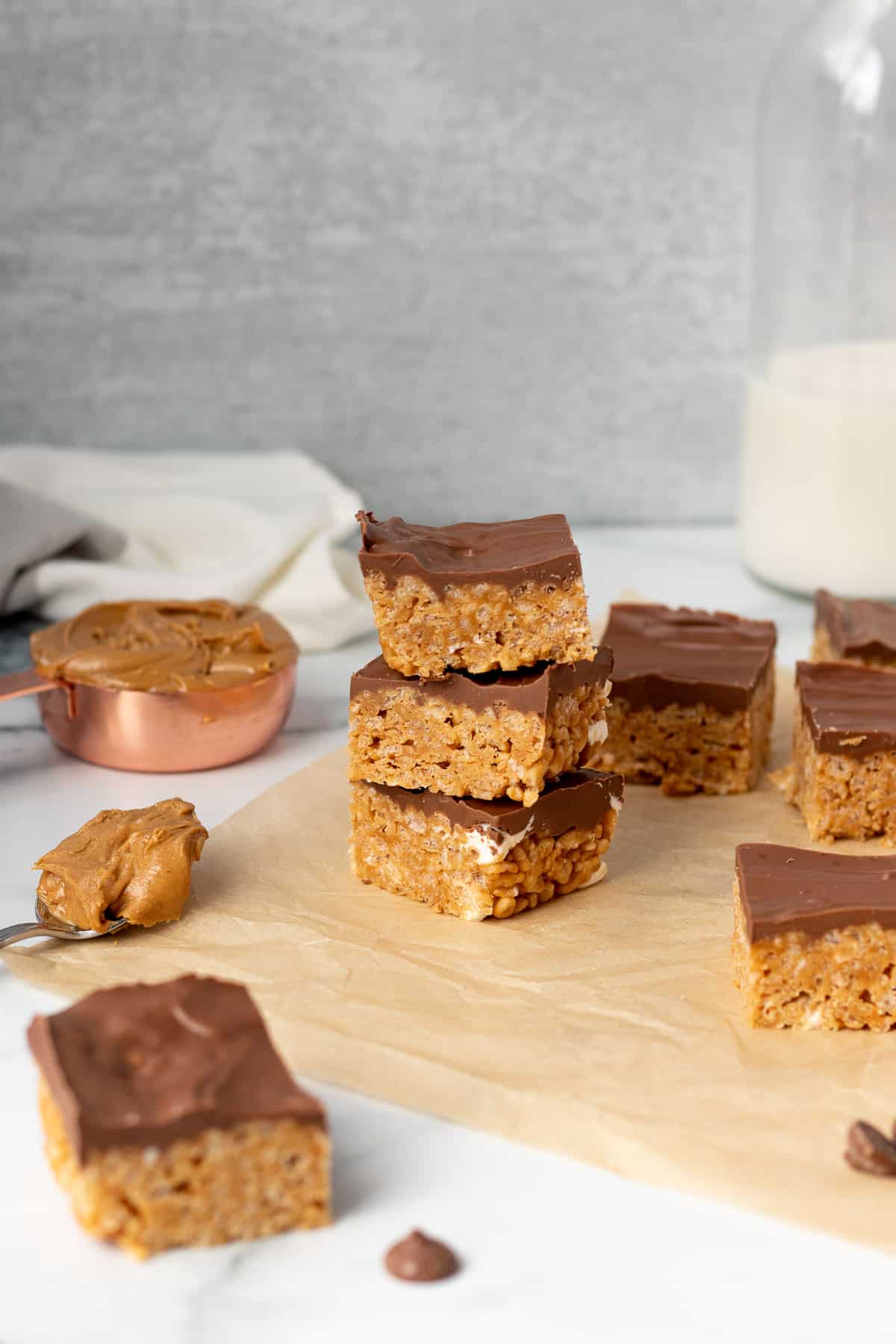 rice krispie treats on brown parchment paper with measuring cup and spoon of peanut butter and glass of milk in background