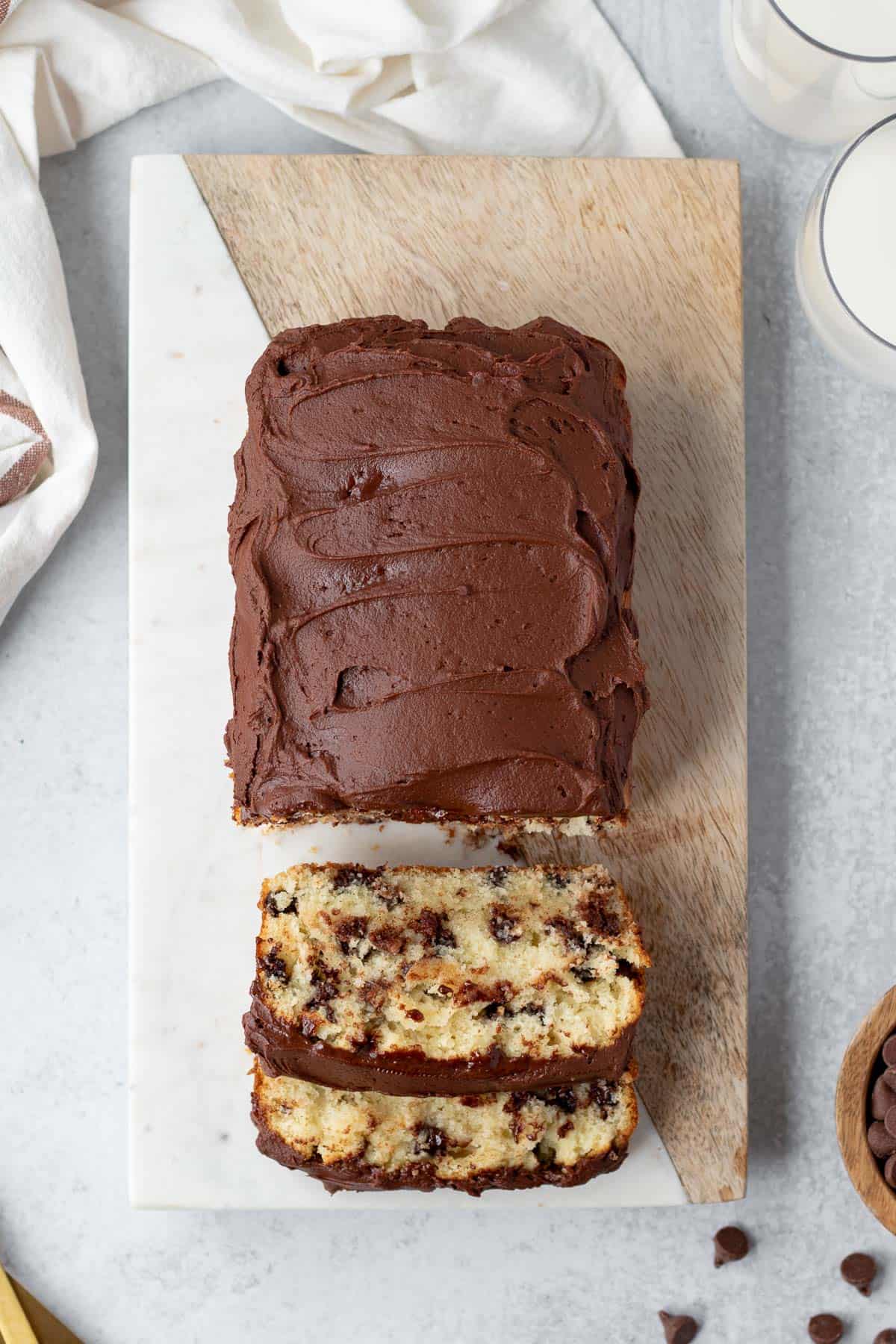 top view of a chocolate chip loaf cake frosted with chocolate frosting on top of a marble and wood cutter board and partially sliced