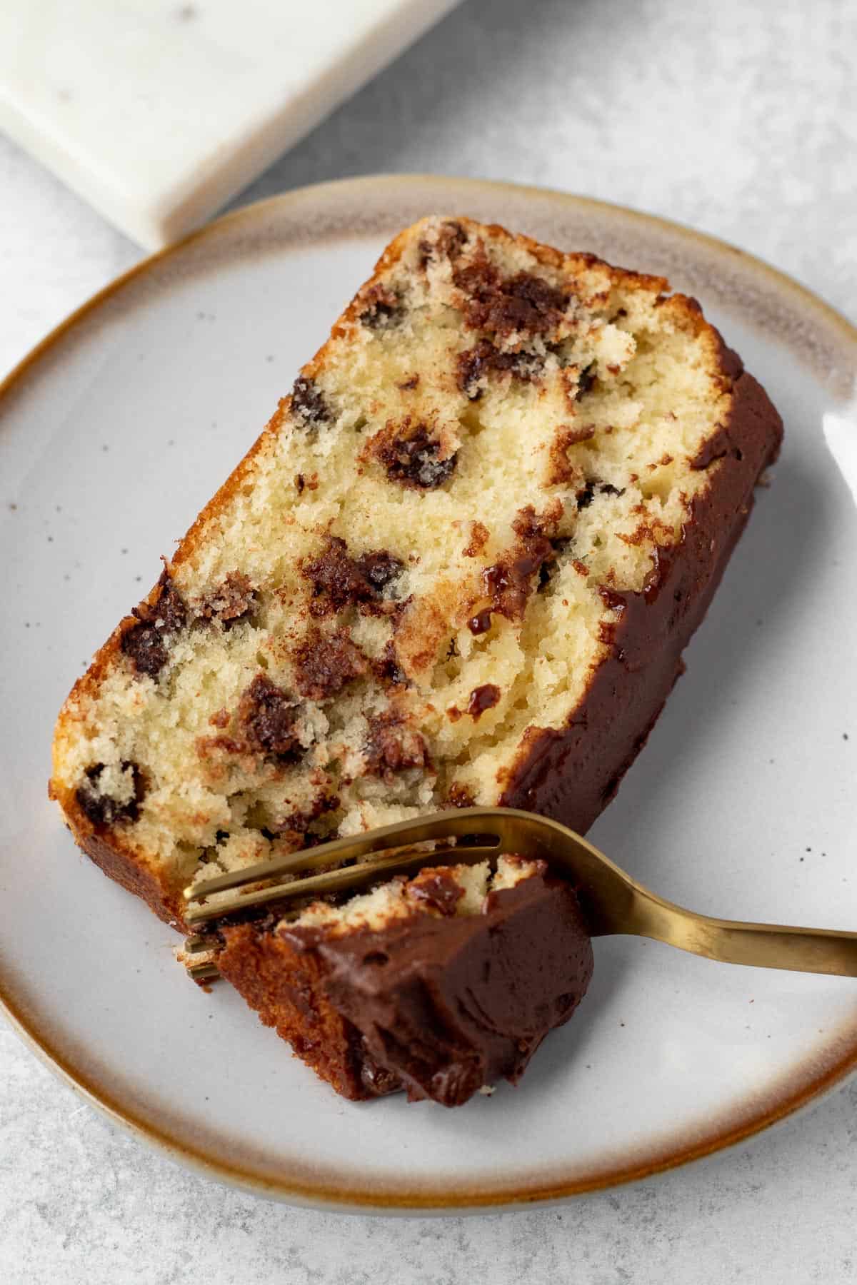 slice of chocolate chip loaf cake on a plate with a gold fork cutting a bite out of it