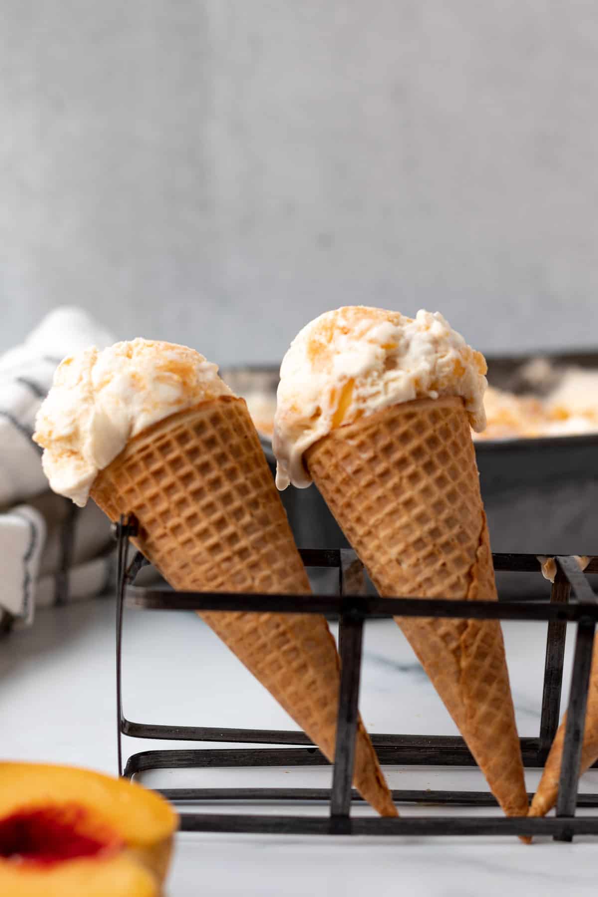 2 ice cream cones standing upright with peach ice cream and a container of ice cream in the background