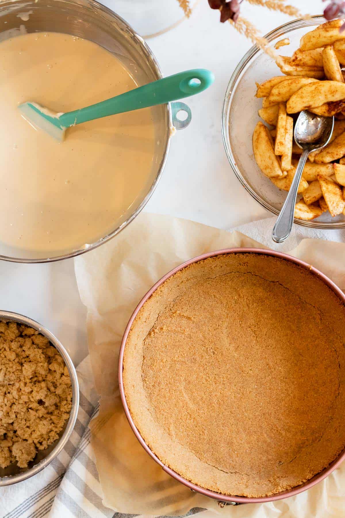 graham cracker crust in a springform pain, bowl of cheesecake filling, bowl of cheesecake filling and bowl of apples waiting to be assembled together