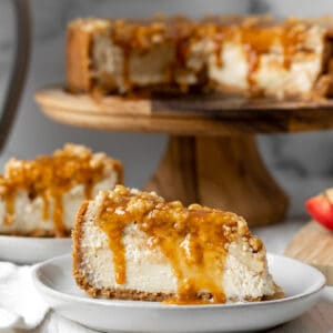 slices of apple crumble cheesecake on white plates with full cheesecake on a wood cake stand in the background