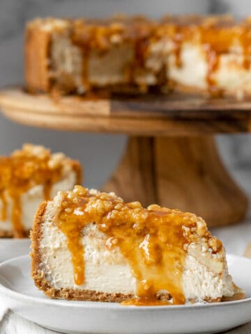 slices of apple crumble cheesecake on white plates with full cheesecake on a wood cake stand in the background