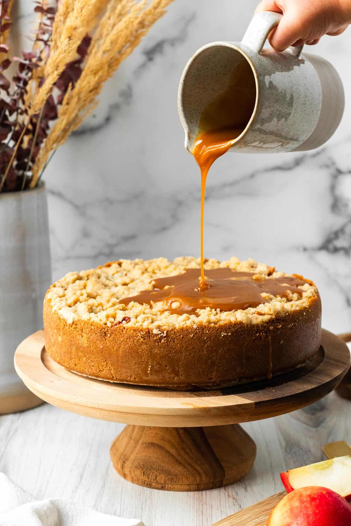 apple crumble cheesecake on a wooden cake stand with a white pitcher pouring caramel on top