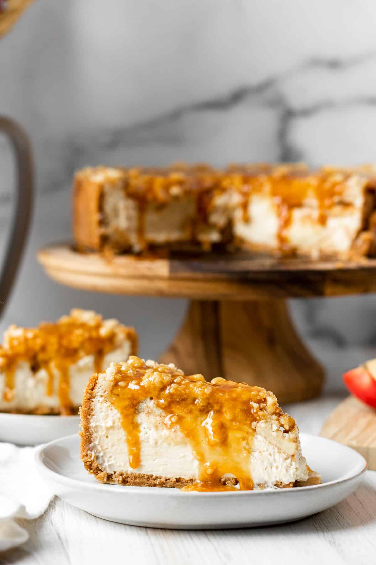 slices of apple crumble cheesecake on white plates with full cheesecake on a wood cake stand in the background