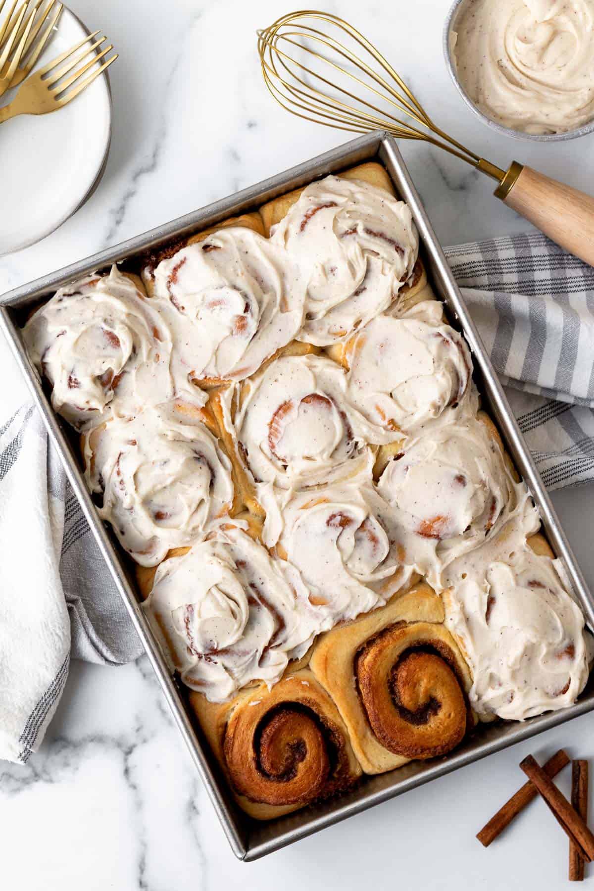 pan of brioche cinnamon rolls frosted except for 2 rolls with a kitchen towel underneath the pan