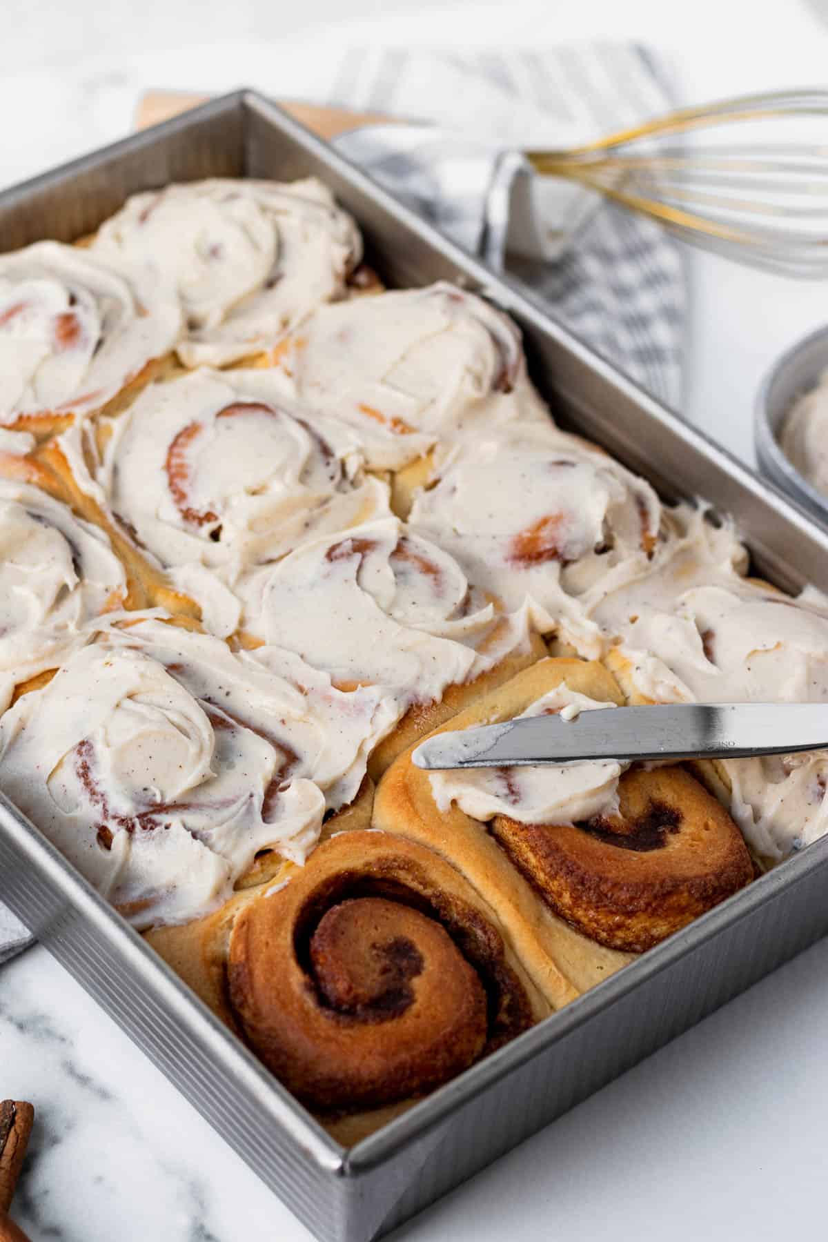 pan of brioche cinnamon rolls partially frosted with a knife spreading frosting