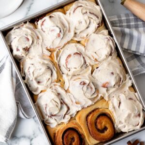 pan of brioche cinnamon rolls frosted except for 2 rolls with a kitchen towel underneath the pan