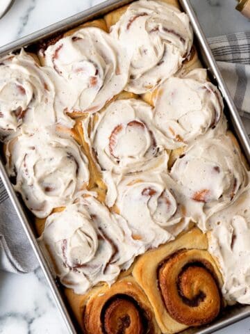 pan of brioche cinnamon rolls frosted except for 2 rolls with a kitchen towel underneath the pan