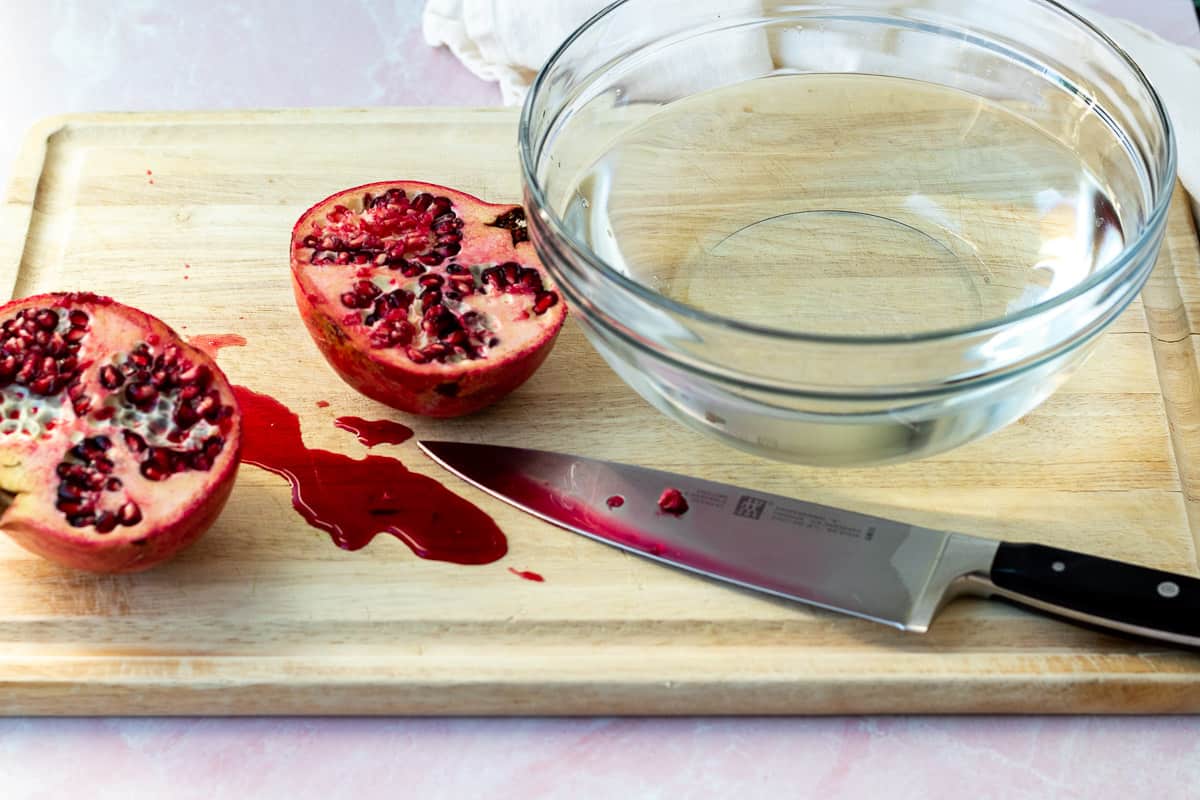 a pomegranate cut in half on a cutting board with a bowl of water next to it