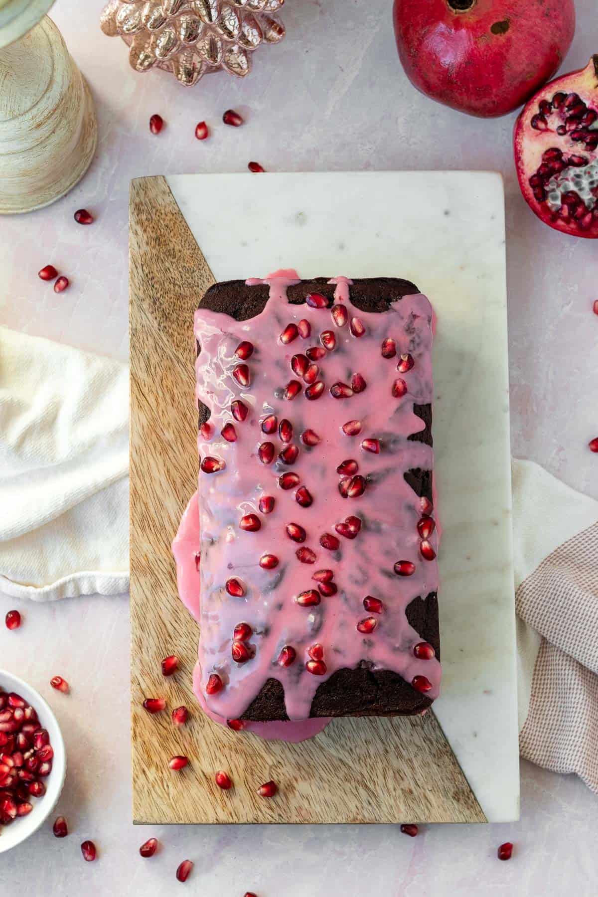 chocolate loaf cake with a pomegranate glaze and pomegranate seeds sitting on top of a wooden and marble cutting boardg