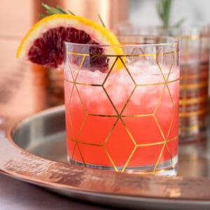 a blood orange gin cocktail in a clear and gold cocktail glass with a garnish of an orange slice and rosemary sprig