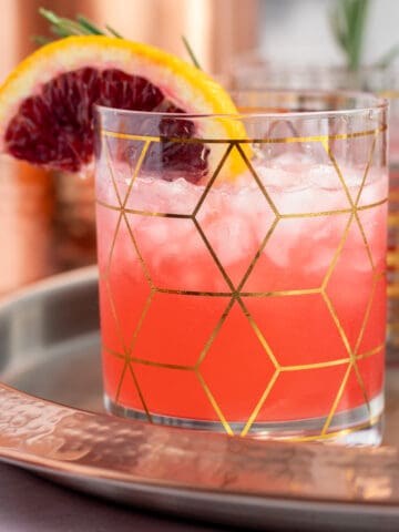 a blood orange gin cocktail in a clear and gold cocktail glass with a garnish of an orange slice and rosemary sprig