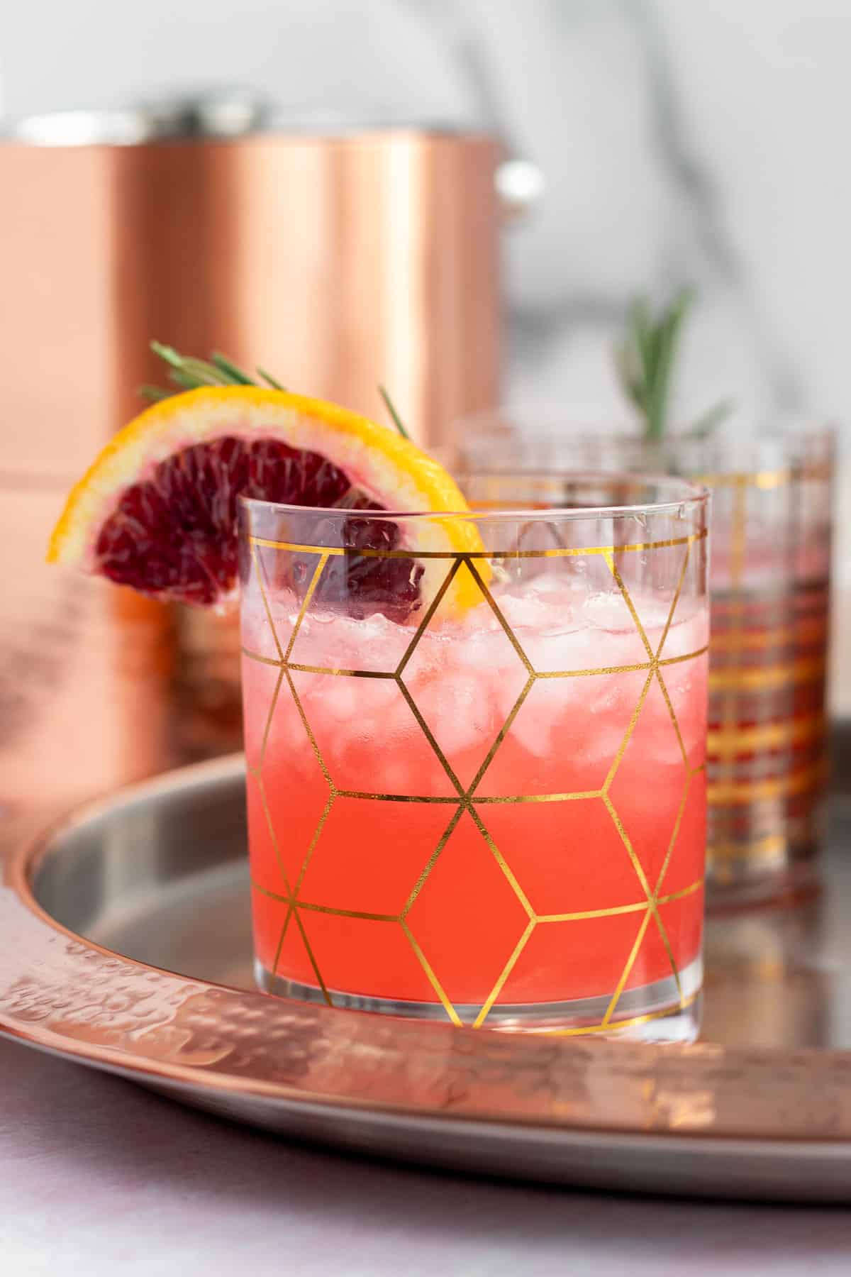 a blood orange gin cocktail in a clear and gold cocktail glass with a garnish of an orange slice and rosemary sprig