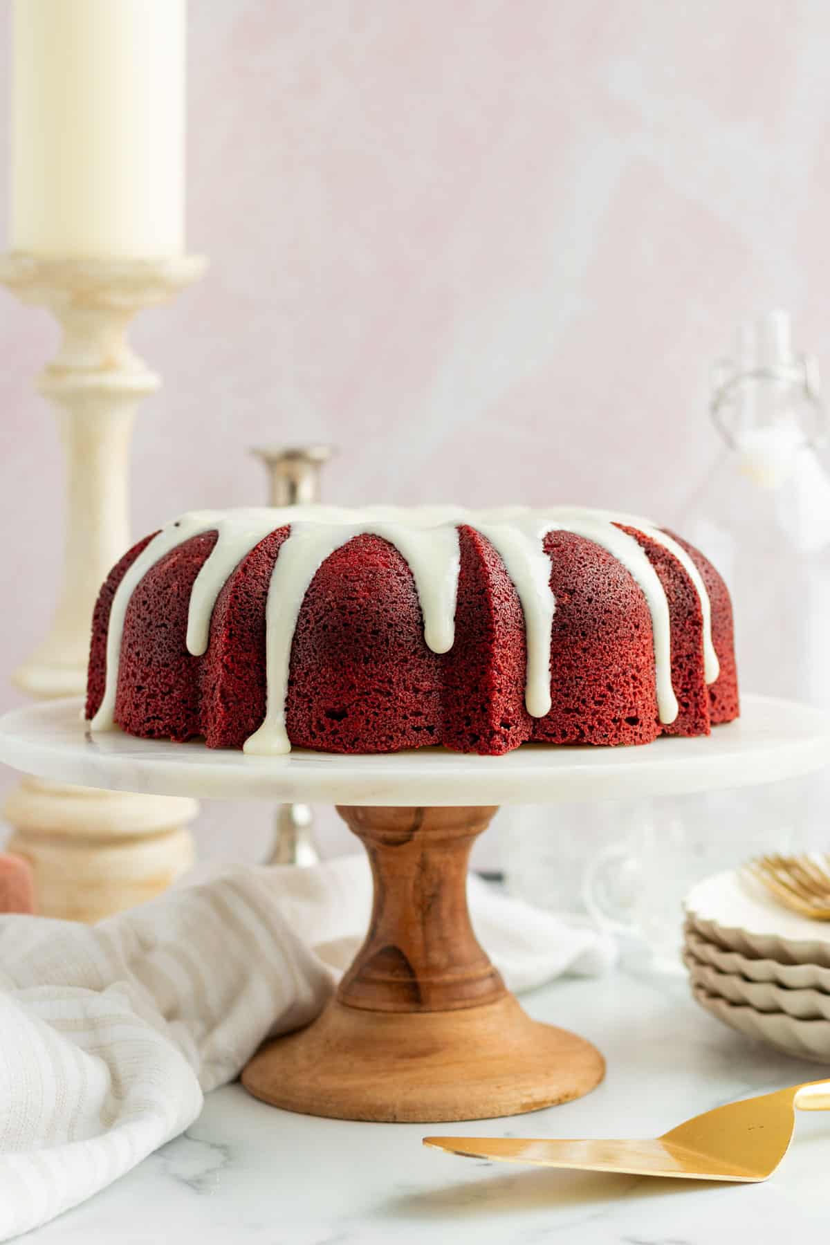 red velvet bundt cake on a marble and wood cake stand with a stack of plates and candlesticks in the background