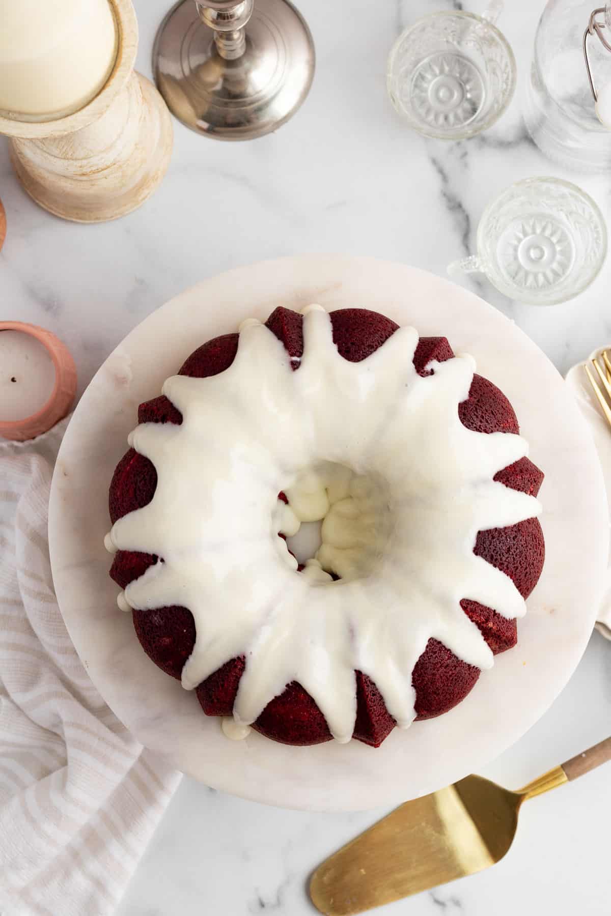 top view of a red velvet bundt cake with a cream cheese glaze sitting on a marble cake stand with a gold cake server nearby