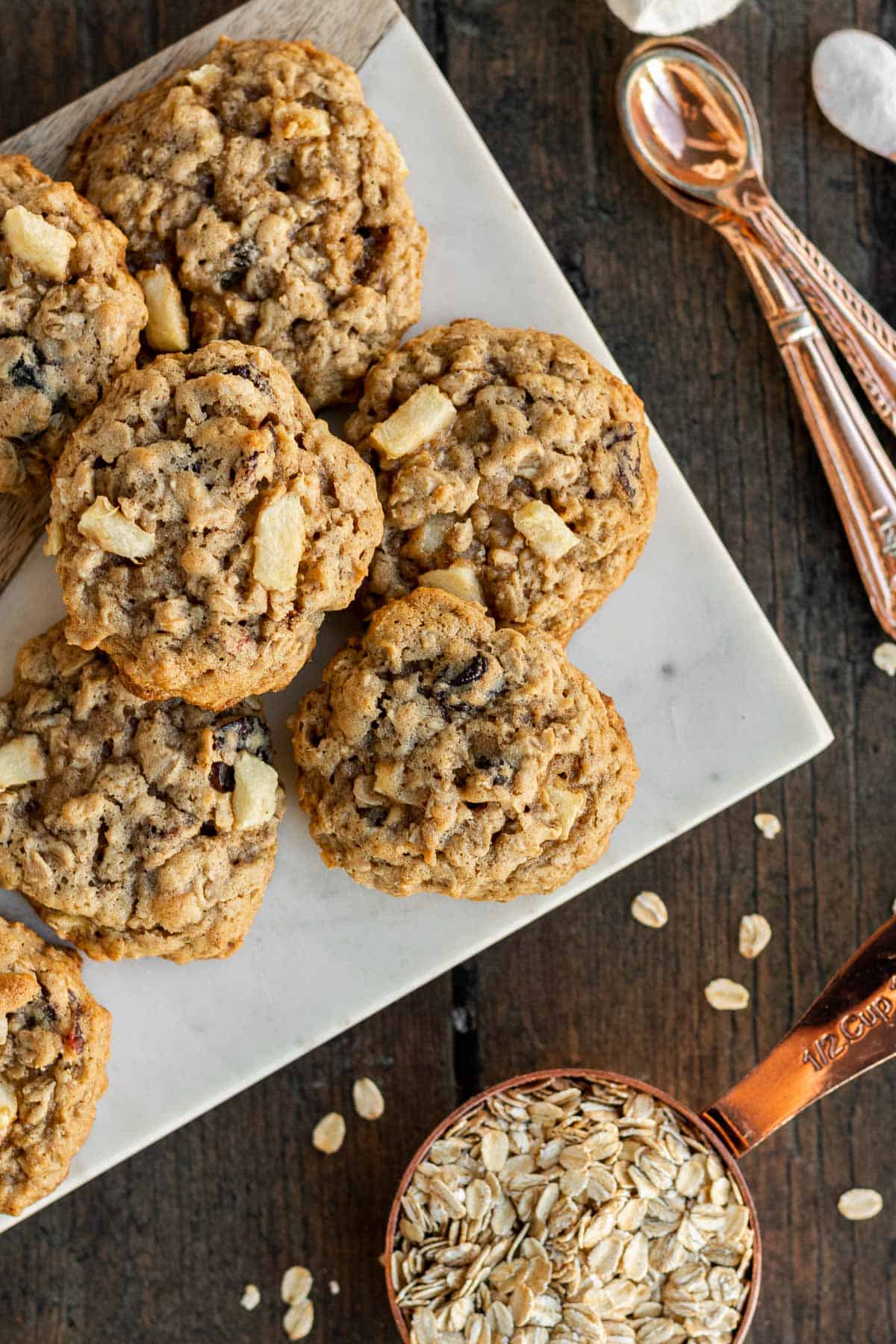 a pile of apple oatmeal cookies on a marble tray with a gold measuring cup of oatmeal sitting near by