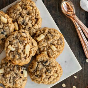 a pile of apple oatmeal cookies on a marble tray with a gold measuring cup of oatmeal sitting near by